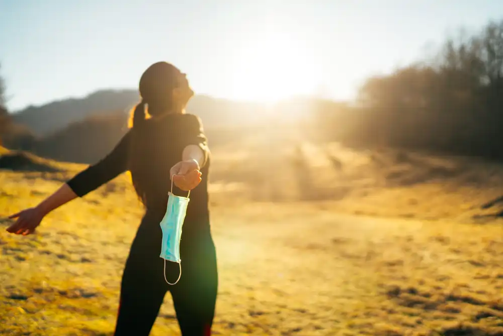 Eine Frau steht im Sonnenlicht und hält eine blaue Flasche in der Hand, wobei ein heller Hintergrund die leuchtenden Farben der Szene hervorhebt.
