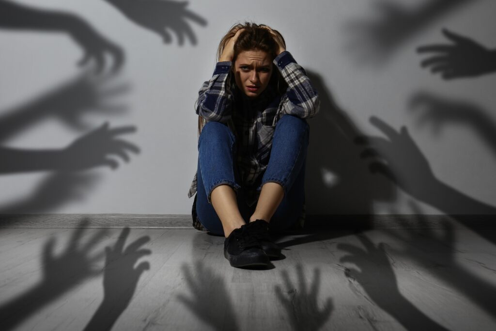 Symbolbild Pranoia - Eine Frau sitzt auf dem Boden und verzweifelt, weil Schatten von Händen nach ihr greifen.